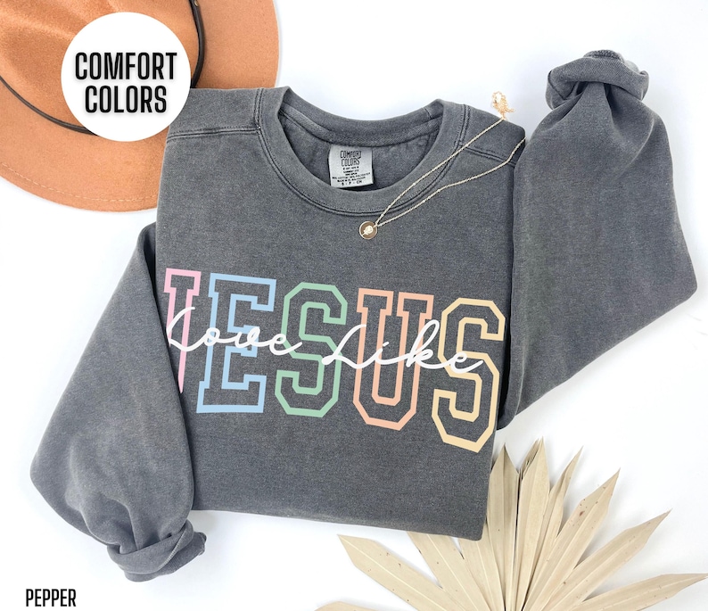 May include: A dark grey sweatshirt with the words "Jesus" in rainbow colours and "love like" in white script underneath. The sweatshirt is made by Comfort Colors.