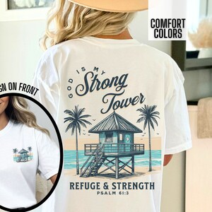 Puede incluir: Camiseta blanca con un gr&aacute;fico de una escena de playa con una torre de salvavidas y palmeras. El texto "God is my Strong Tower" est&aacute; escrito en una fuente cursiva sobre la torre. El texto "Refuge & Strength Psalm 61:3" est&aacute; escrito debajo de la torre.
