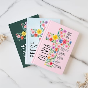 May include: Three journals with floral cross designs. One is dark green with a floral cluster, another is light blue with the name "PFFSE", and the third is pink with the name "OLIVIA" and inspirational words.