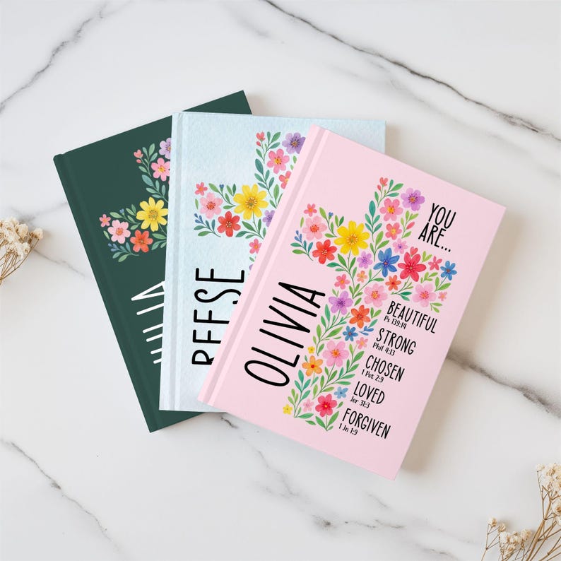 May include: Three journals with floral cross designs. One is dark green with a floral cluster, another is light blue with the name "PFFSE", and the third is pink with the name "OLIVIA" and inspirational words.