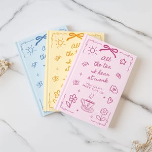 K&ouml;nnte beinhalten: Drei Notizb&uuml;cher in Pastellfarben: Blau, Gelb und Rosa. Jedes Notizbuch hat einen dekorativen Rand mit einer Schleife, Sonne, Schmetterlingen und Text. Auf dem rosa Notizbuch steht: "All the tea I hear at work. You can't make this up."