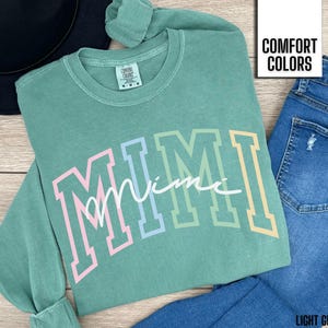 May include: A light green sweatshirt with the word "MIMI" in large, colorful letters. The word "Mimi" is in a script font. The sweatshirt is paired with blue jeans and a black hat. The text "COMFORT COLORS" is in a white box.