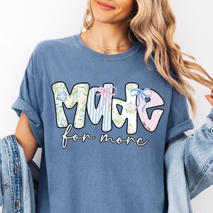 Op de afbeelding: Een blauw t-shirt met de tekst "MADE for more" in een speels lettertype. De letters zijn versierd met bloemen-, gestreepte en ruitpatronen. Het shirt heeft een losse pasvorm en opgerolde mouwen.