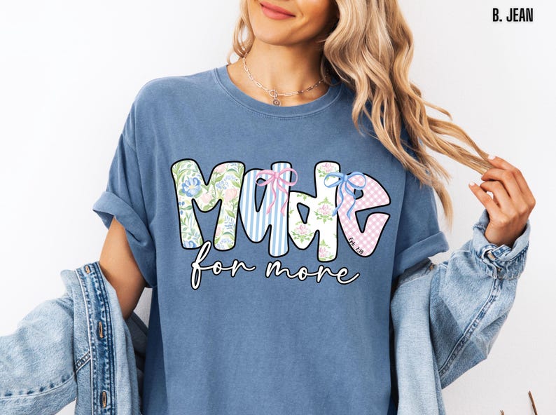 Op de afbeelding: Een blauw t-shirt met de tekst "MADE for more" in een speels lettertype. De letters zijn versierd met bloemen-, gestreepte en ruitpatronen. Het shirt heeft een losse pasvorm en opgerolde mouwen.