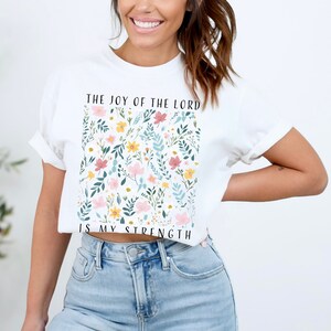 Op de afbeelding: Wit kort T-shirt met een bloemenpatroon en de tekst "The Joy of the Lord is My Strength".