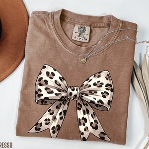 Puede incluir: Una camiseta marr&oacute;n con un gran lazo de estampado de leopardo en la parte delantera. La camiseta est&aacute; hecha de un material suave y c&oacute;modo y tiene un aspecto desgastado.