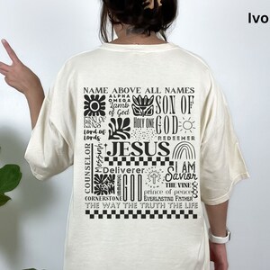 May include: Ivory t-shirt with a black graphic print of the word "Jesus" surrounded by other religious phrases and symbols. The text includes "Name Above All Names", "Son of God", "Holy One", "Redeemer", "I Am Savior", "The Vine", "Prince of Peace", "Everlasting Father", "The Way The Truth The Life", "Alpha Omega", "King of Kings", "Lord of Lords", "Counselor", "Messiah", "Deliverer", "Cornerstone", and "Immanuel God".