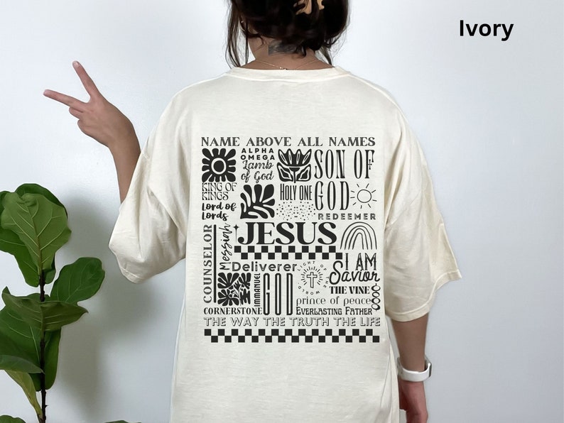 May include: Ivory t-shirt with a black graphic print of the word "Jesus" surrounded by other religious phrases and symbols. The text includes "Name Above All Names", "Son of God", "Holy One", "Redeemer", "I Am Savior", "The Vine", "Prince of Peace", "Everlasting Father", "The Way The Truth The Life", "Alpha Omega", "King of Kings", "Lord of Lords", "Counselor", "Messiah", "Deliverer", "Cornerstone", and "Immanuel God".