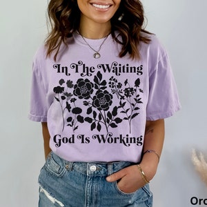 Op de afbeelding: Een lavendelkleurig T-shirt met een zwart bloemenontwerp en de tekst "In The Waiting God Is Working".