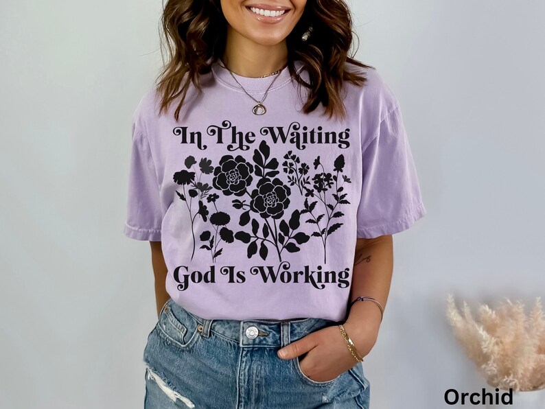 Op de afbeelding: Een lavendelkleurig T-shirt met een zwart bloemenontwerp en de tekst "In The Waiting God Is Working".