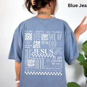 May include: A blue jean t-shirt with a white graphic print of the word "Jesus" surrounded by other religious phrases and symbols. The text includes "Name Above All Names", "Son of God", "Holy One", "Redeemer", "Counselor", "Messiah", "Deliverer", "I Am Savior", "The Vine", "Prince of Peace", "Everlasting Father", "The Way The Truth The Life", and "Cornerstone".