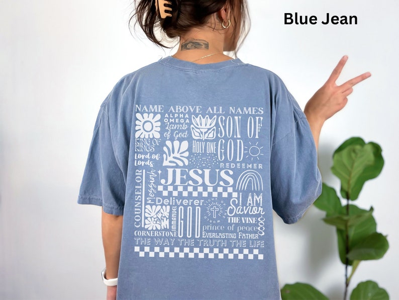 May include: A blue jean t-shirt with a white graphic print of the word "Jesus" surrounded by other religious phrases and symbols. The text includes "Name Above All Names", "Son of God", "Holy One", "Redeemer", "Counselor", "Messiah", "Deliverer", "I Am Savior", "The Vine", "Prince of Peace", "Everlasting Father", "The Way The Truth The Life", and "Cornerstone".