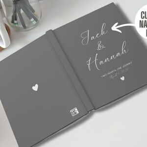 K&ouml;nnte beinhalten: Graues Hardcover-G&auml;stebuch mit wei&szlig;em Text, der "Jack & Hannah Zwei Herzen, eine Reise 4. April 2020" lautet.