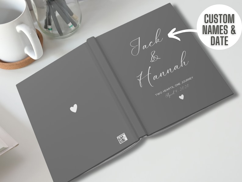 K&ouml;nnte beinhalten: Graues Hardcover-G&auml;stebuch mit wei&szlig;em Text, der "Jack & Hannah Zwei Herzen, eine Reise 4. April 2020" lautet.