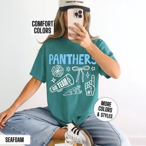 Puede incluir: Una camiseta de manga corta color verde azulado con el texto azul claro PANTHERS e ilustraciones gr&aacute;ficas blancas de un pomp&oacute;n, un lazo, un meg&aacute;fono con el texto GO TEAM, un bander&iacute;n y un dedo de espuma con el n&uacute;mero uno.
