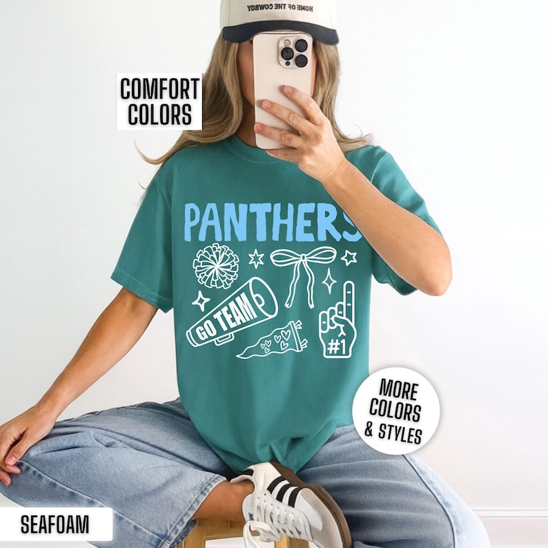 Puede incluir: Una camiseta de manga corta color verde azulado con el texto azul claro PANTHERS e ilustraciones gr&aacute;ficas blancas de un pomp&oacute;n, un lazo, un meg&aacute;fono con el texto GO TEAM, un bander&iacute;n y un dedo de espuma con el n&uacute;mero uno.
