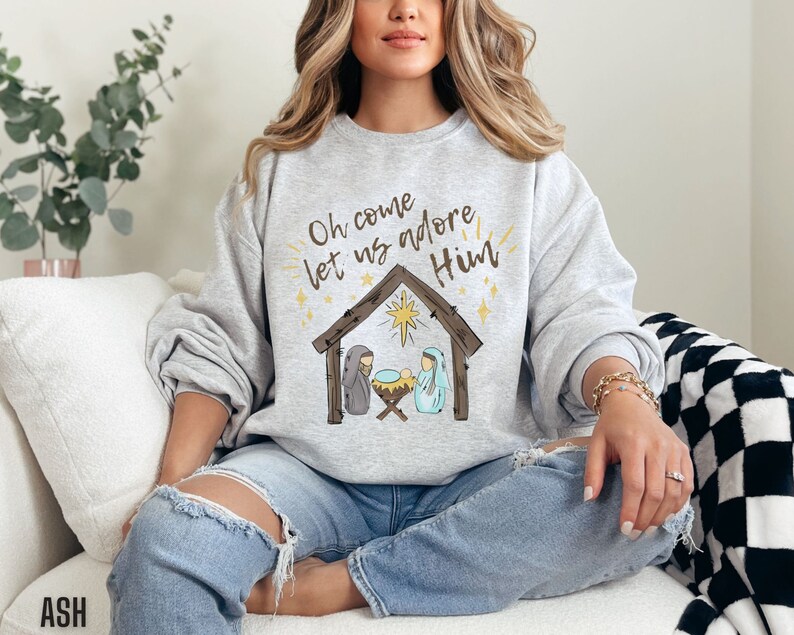 Puede incluir: Una sudadera gris con el texto "Oh come let us adore Him" y una representaci&oacute;n de la escena de la natividad.