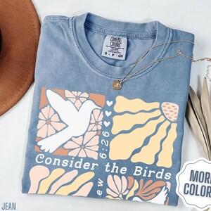 Op de afbeelding: Een lichtblauw T-shirt met een grafisch ontwerp met een witte vogel, bloemen en de tekst "Consider the Birds 6:26".