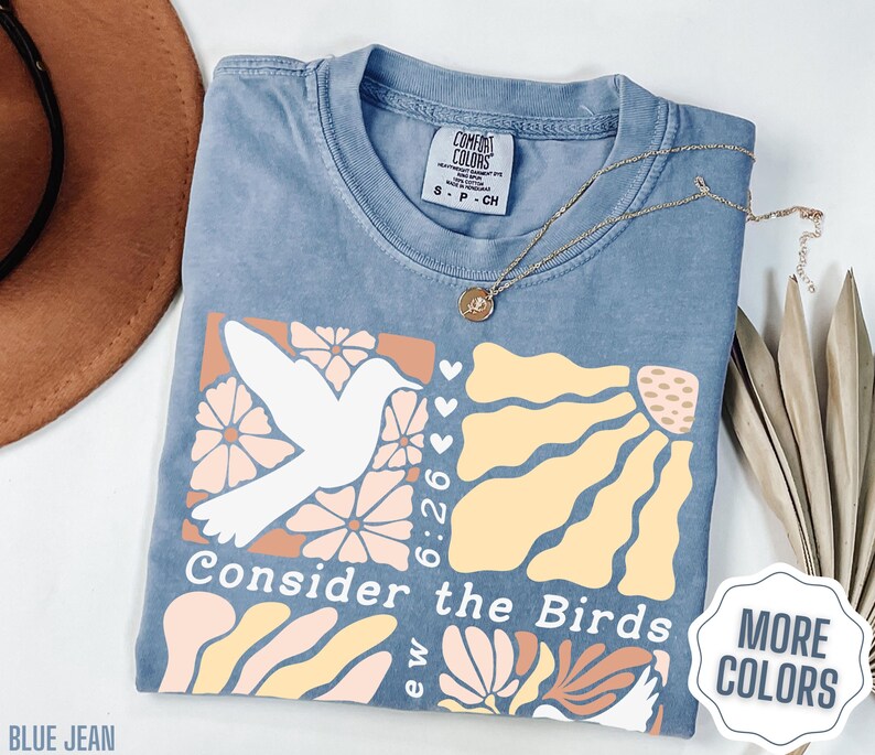 Op de afbeelding: Een lichtblauw T-shirt met een grafisch ontwerp met een witte vogel, bloemen en de tekst "Consider the Birds 6:26".