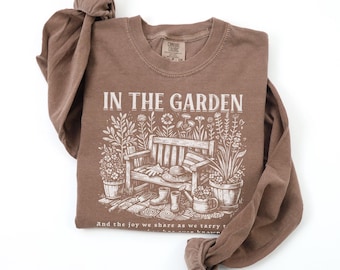 May include: A brown long-sleeve t-shirt with the text "In the Garden" and a graphic of a bench, gardening tools, and plants. The text below the graphic reads "And the joy we share as we tarry to see green grow."