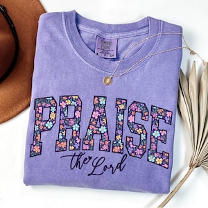 May include: A violet-colored t-shirt with the word "PRAISE" in large, floral-patterned letters, and "the Lord" in script below. A gold necklace and a brown hat are visible in the background.