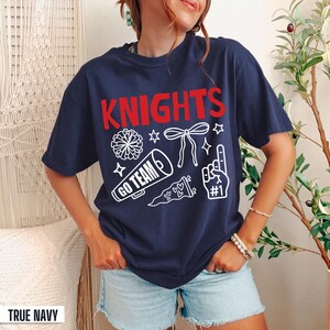 Puede incluir: Una camiseta de manga corta azul marino con el texto rojo KNIGHTS y gr&aacute;ficos blancos de un pomp&oacute;n, un lazo, un meg&aacute;fono con el texto GO TEAM, un bander&iacute;n y un dedo de espuma con el texto #1 impresos en la parte frontal del pecho.