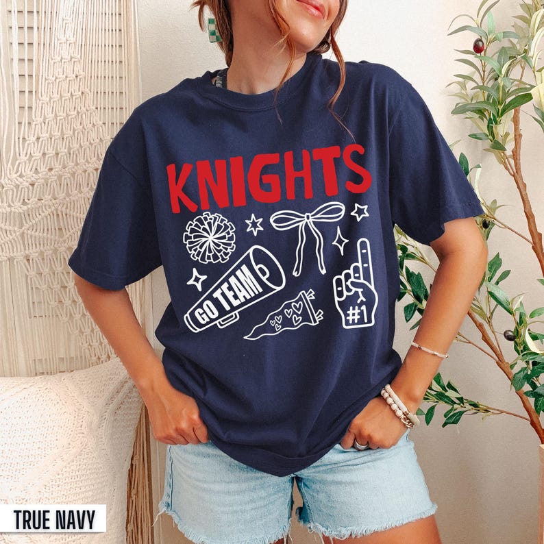 Puede incluir: Una camiseta de manga corta azul marino con el texto rojo KNIGHTS y gr&aacute;ficos blancos de un pomp&oacute;n, un lazo, un meg&aacute;fono con el texto GO TEAM, un bander&iacute;n y un dedo de espuma con el texto #1 impresos en la parte frontal del pecho.