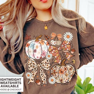 Puede incluir: Sudadera marr&oacute;n con un dise&ntilde;o floral de calabaza, margaritas, lazos y las palabras "Autumn Vibes", "Cozy", "Falling Leaves" y "Harvest". Tambi&eacute;n se ve el texto "Lightweight Sweatshirts Available".