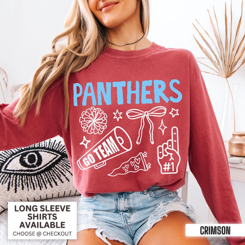 Puede incluir: Una sudadera de manga larga color carmes&iacute; con el texto azul claro PANTHERS e ilustraciones gr&aacute;ficas blancas de un pomp&oacute;n, un lazo, un meg&aacute;fono con el texto GO TEAM, un bander&iacute;n con corazones y un dedo de espuma con el texto #1.