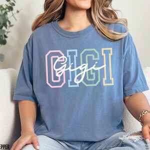 May include: A blue oversized t-shirt with the word "Gigi" in colorful block letters and a white cursive script.