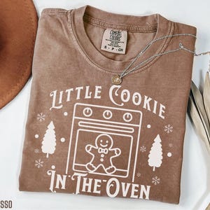 May include: Espresso-colored t-shirt with the text "Little Cookie In The Oven" in white. The design includes a gingerbread man in an oven, snowflakes, and Christmas trees. A silver necklace rests on the shirt.