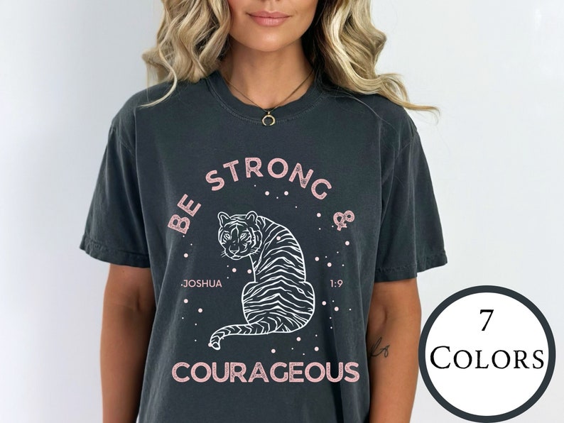 May include: A dark gray t-shirt with a white tiger graphic and the text "Be Strong & Courageous" in pink lettering. The text "Joshua 1:9" is also included in white lettering.