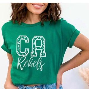 May include: A green t-shirt with the text "CA Rebels" in white. The letters "CA" are in a block letter style with a leopard print pattern. The word "Rebels" is in a cursive font.