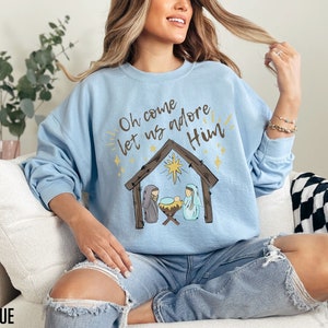 Puede incluir: Una sudadera de color azul claro con un gr&aacute;fico de la natividad de Navidad y el texto "Oh come let us adore Him".