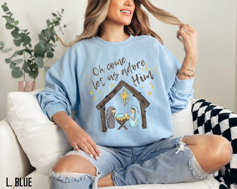 Puede incluir: Una sudadera de color azul claro con un gr&aacute;fico de la natividad de Navidad y el texto "Oh come let us adore Him".