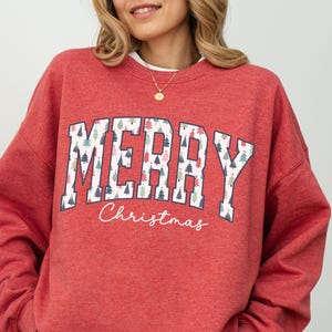 May include: Red Christmas sweatshirt with a white "Merry Christmas" graphic featuring a pattern of small Christmas trees.