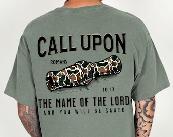 May include: A green t-shirt with a camouflage duck call design. The text "CALL UPON" is printed in black above the duck call. The text "ROMANS 10:13 THE NAME OF THE LORD AND YOU WILL BE SAVED" is printed in black below the duck call.