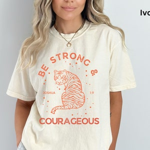 May include: Ivory t-shirt with a graphic of a tiger and the text "Be Strong & Courageous". The graphic also includes the text "Joshua 1:9".