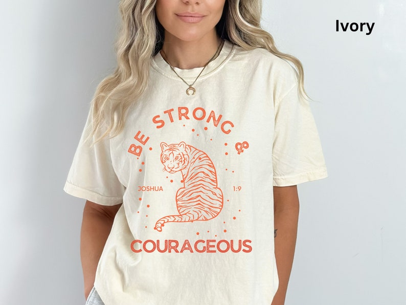 May include: Ivory t-shirt with a graphic of a tiger and the text "Be Strong & Courageous". The graphic also includes the text "Joshua 1:9".