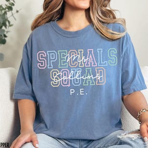 May include: A blue t-shirt featuring the text "SPECIALS SQUAD" in colourful block letters and the name "Mrs. Collins" in cursive. The letters "P.E." are below. The shirt is a relaxed, casual style.