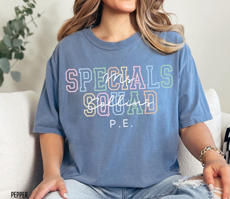 May include: A blue t-shirt featuring the text "SPECIALS SQUAD" in colourful block letters and the name "Mrs. Collins" in cursive. The letters "P.E." are below. The shirt is a relaxed, casual style.