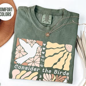 Op de afbeelding: Een groen T-shirt met een wit duifje en een bloemenontwerp. De tekst "Consider the Birds" staat op het shirt gedrukt. Het shirt is gemaakt van een zacht, comfortabel materiaal.