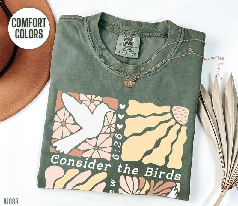 Op de afbeelding: Een groen T-shirt met een wit duifje en een bloemenontwerp. De tekst "Consider the Birds" staat op het shirt gedrukt. Het shirt is gemaakt van een zacht, comfortabel materiaal.