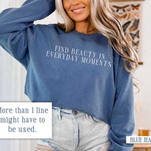 May include: A blue cropped sweatshirt with the text "FIND BEAUTY IN EVERYDAY MOMENTS" printed in white on the front. The sweatshirt is being worn by a person with long blonde hair and is paired with light blue denim shorts.