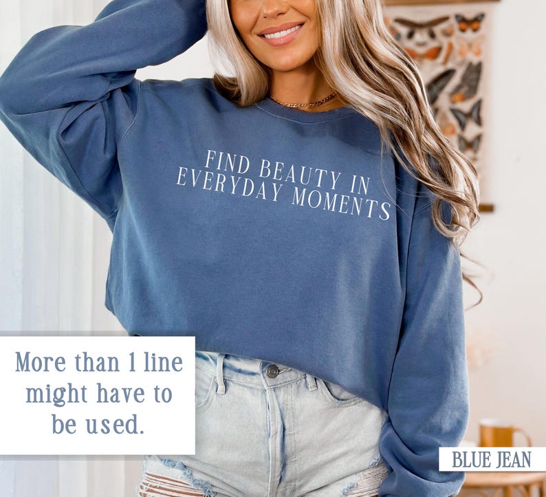 May include: A blue cropped sweatshirt with the text "FIND BEAUTY IN EVERYDAY MOMENTS" printed in white on the front. The sweatshirt is being worn by a person with long blonde hair and is paired with light blue denim shorts.