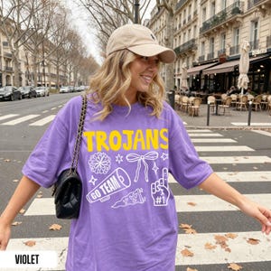 Puede incluir: Una camiseta violeta de manga corta con el texto amarillo TROJANS y gr&aacute;ficos de l&iacute;neas blancas que incluyen un pomp&oacute;n, un lazo, un meg&aacute;fono con el texto GO TEAM, un bander&iacute;n con corazones y un dedo de espuma con el texto #1.
