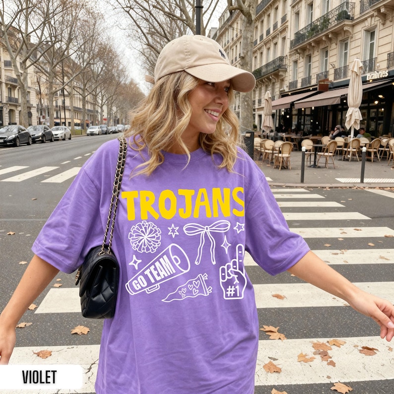 Puede incluir: Una camiseta violeta de manga corta con el texto amarillo TROJANS y gr&aacute;ficos de l&iacute;neas blancas que incluyen un pomp&oacute;n, un lazo, un meg&aacute;fono con el texto GO TEAM, un bander&iacute;n con corazones y un dedo de espuma con el texto #1.