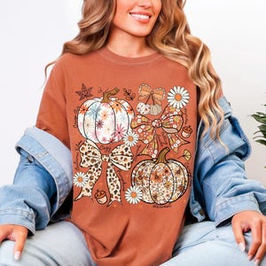 Puede incluir: Camiseta de color &oacute;xido con un gr&aacute;fico tem&aacute;tico de oto&ntilde;o. El dise&ntilde;o incluye calabazas, lazos y elementos florales. Tambi&eacute;n son visibles las palabras "Autumn Vibes", "Cozy" y "Harvest". La camiseta est&aacute; hecha de un material suave.
