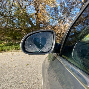 Op de afbeelding: Een zijspiegel van een auto met een sticker van een gebroken hart. De spiegel is bedekt met condensatie druppels.