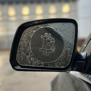 May include: A car side mirror with a Bitcoin symbol sticker on it. The mirror is covered in water droplets.